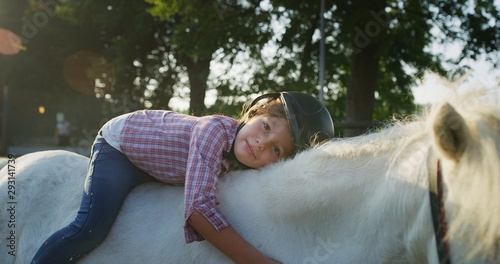 Принти на полотні Authentic shot of a cute little girl with a jockey helmet is lying on a white pony horse and caressing her at riding stable with a sunshine