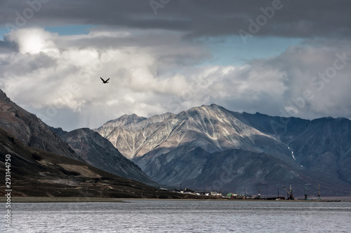 Epic landscape with a small port town on the coast of the Bering Sea. Majestic mountains and clouds. In the distance the cranes of the seaport. Location place: Egvekinot, Chukotka, Far East of Russia.