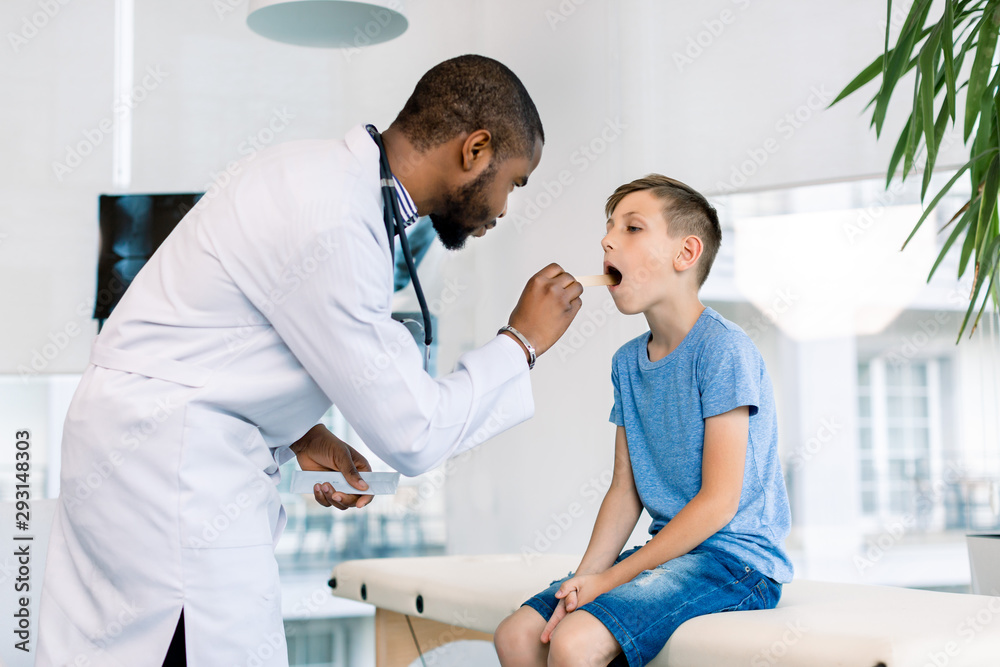 Fototapeta premium Showing throat. Little boy opening mouth showing her throat while visiting young handsome African man doctor in his medical office