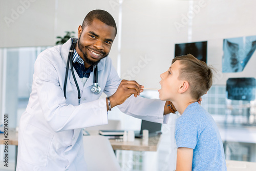 Happy African American male doctor watching patient boy with sore throat in clinic. The doctor examining little boy s throat at hospital
