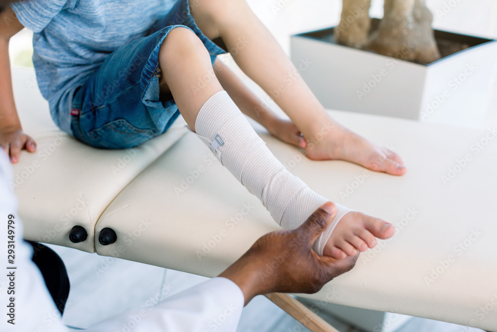 Obraz premium cropped shot of african american doctor putting on bandage on patients injured leg. Doctor first aid, child patient's leg sprain with elastic bandage.