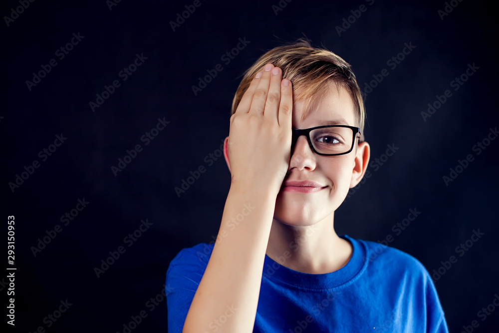A portrait of a boy with eyeglasses close one eye with palm to chech vision in front of dark background. Children, ophthalmology and healthcare concept A portrait of a boy with eyeglasses close one eye with palm to chech vision in front of dark background. Children, ophthalmology and healthcare concept