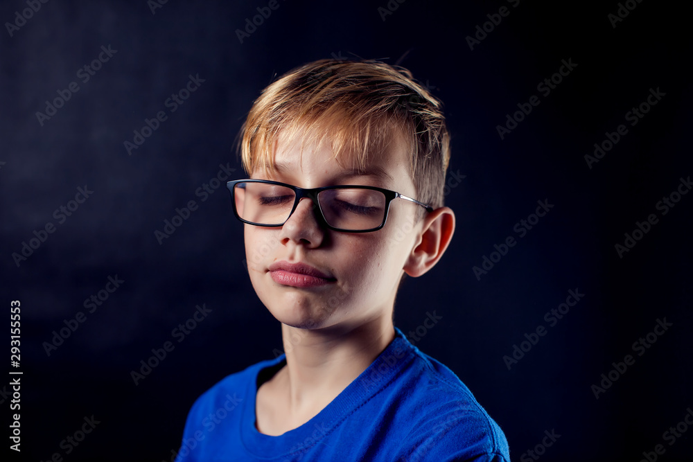 A portrait of a boy with eyeglasses and closed eyes in front of dark background.