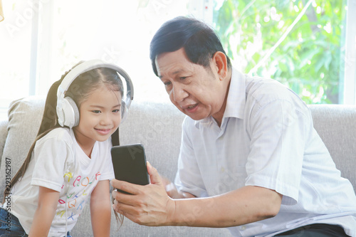 Grandfather is happy playing mobile smartphone with granddaughter. Children wear white headphones interested mobile smartphone in holding hand grandpa.