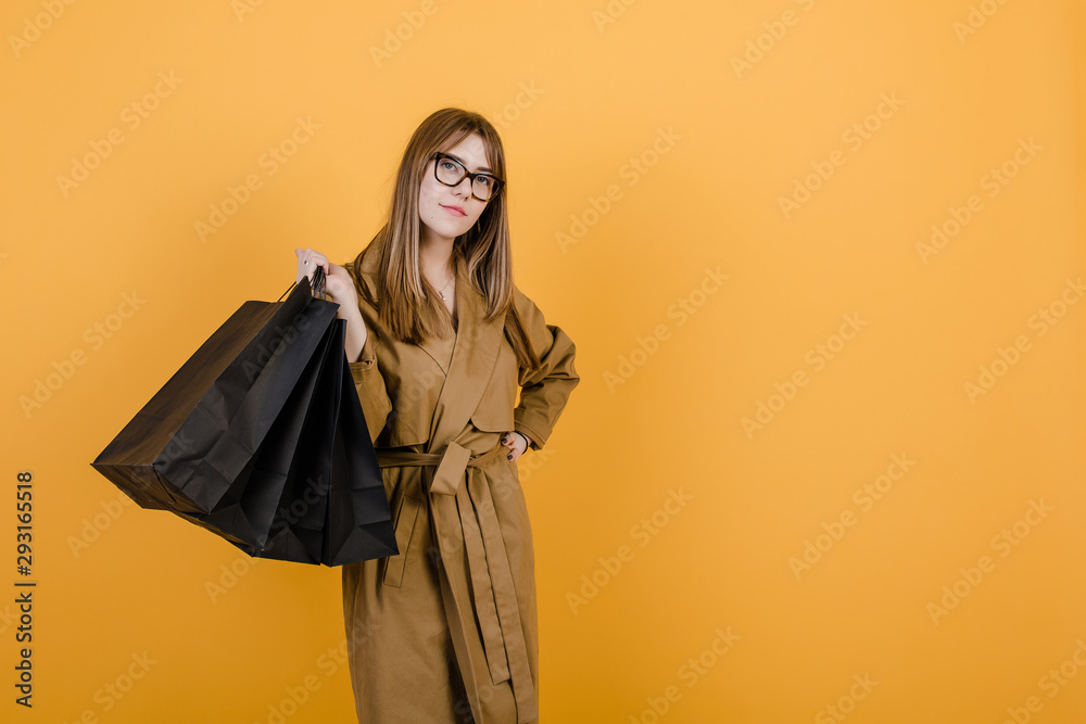 hipster young woman in glasses and fall trench coat with black shopping bags isolated over yellow