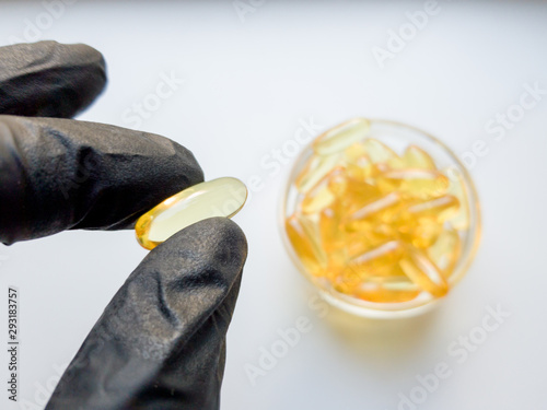 fish oil capsules in hand in a black medical glove on a white background, the hand takes one tablet from a saucer or plate. Hand holding omega 3 capsule on a white background. Healthcare concept