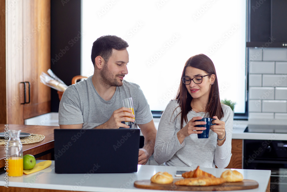 Couple with laptop in pijamas sitting in the kitchen in the morning with pastry and orange juice on table Couple with laptop in pijamas sitting in the kitchen in the morning with pastry and orange juice on table