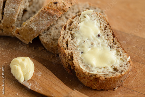 Fényképezés Closeup of buttered slice of whole wheat rustic bread next to cut bread and wood knife