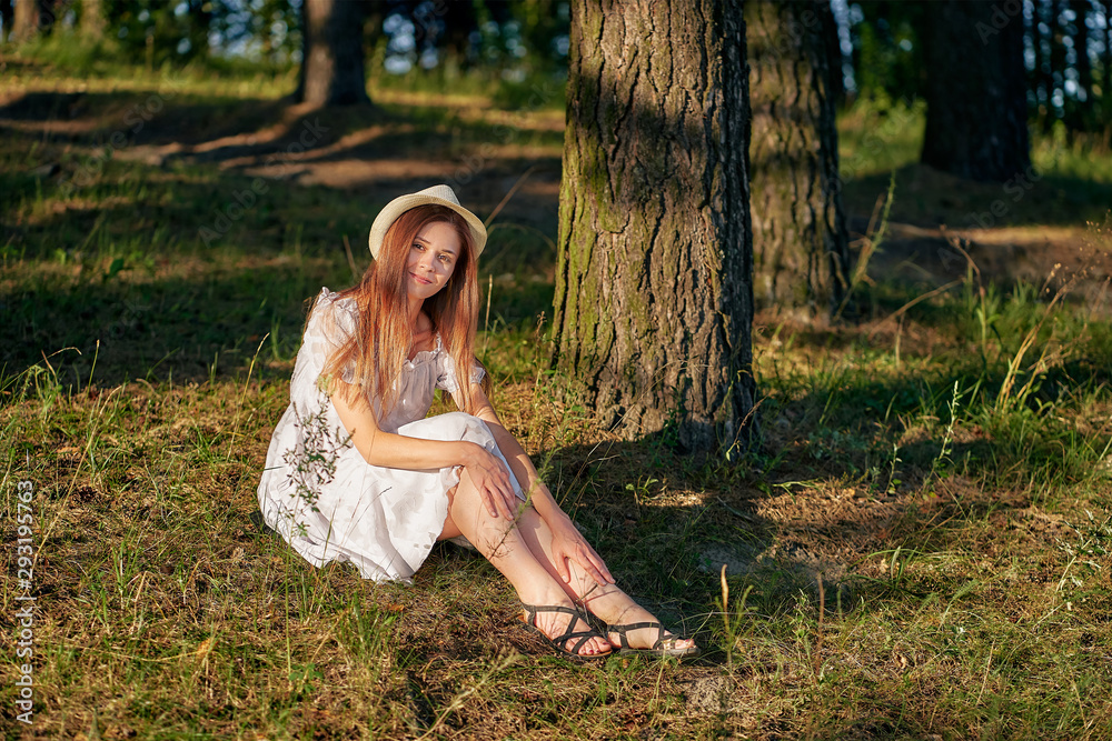 young woman in a summer dress and hat sits under a tree in the forest. copy space