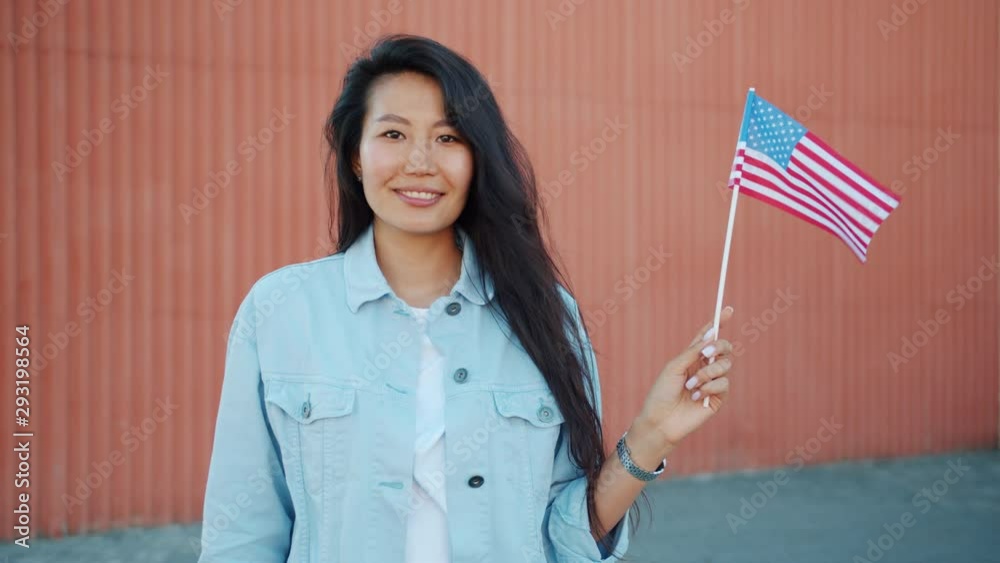 Portrait of pretty Asian girl holding official US flag outdoors smiling looking at camera expressing patriotic feelings. People, countries and tourism concept. Portrait of pretty Asian girl holding official US flag outdoors smiling looking at camera expressing patriotic feelings. People, countries and tourism concept.