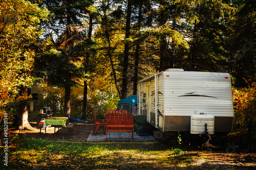 A deserted camper trailer and tent with wooden chairs on outdoor carpet and cloth covered table surrounded by tall pine trees parked in a deserted campsite in a autumn daytime landscape