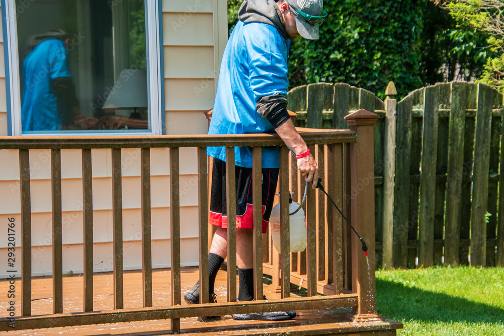 Caucasian young man wearing blue raincoat spraying cleaning detergent on the railing and post of a brown wooden deck in a backyard Caucasian young man wearing blue raincoat spraying cleaning detergent on the railing and post of a brown wooden deck in a backyard