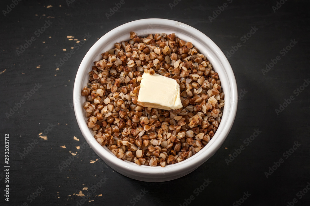 Cooked buckwheat porridge in a deep plate with a slice of butter on a black wooden background. Cooked buckwheat porridge in a deep plate with a slice of butter on a black wooden background.