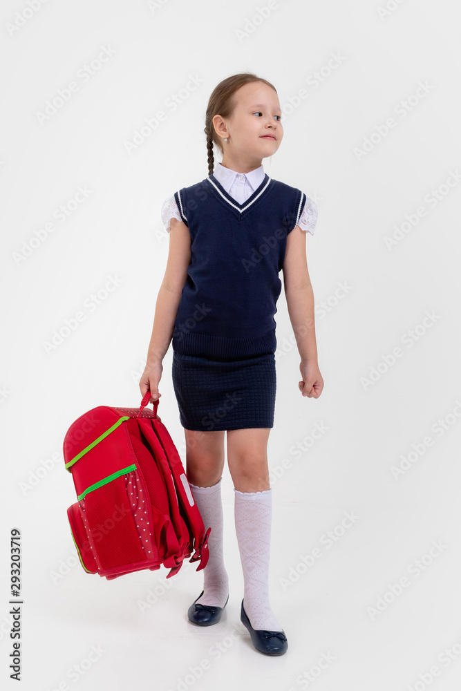 Beautiful small girl schoolgirl in black skirt and blouse goes to school. Behind the girl a big red schoolbag , holding her stack of books. Close-up.Isolated on white background. Beautiful small girl schoolgirl in black skirt and blouse goes to school. Behind the girl a big red schoolbag , holding her stack of books. Close-up.Isolated on white background.