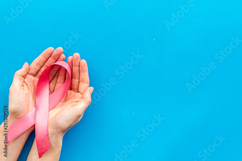 Pink ribbon in hands as symbol of breast cancer awareness on blue background top view copy space