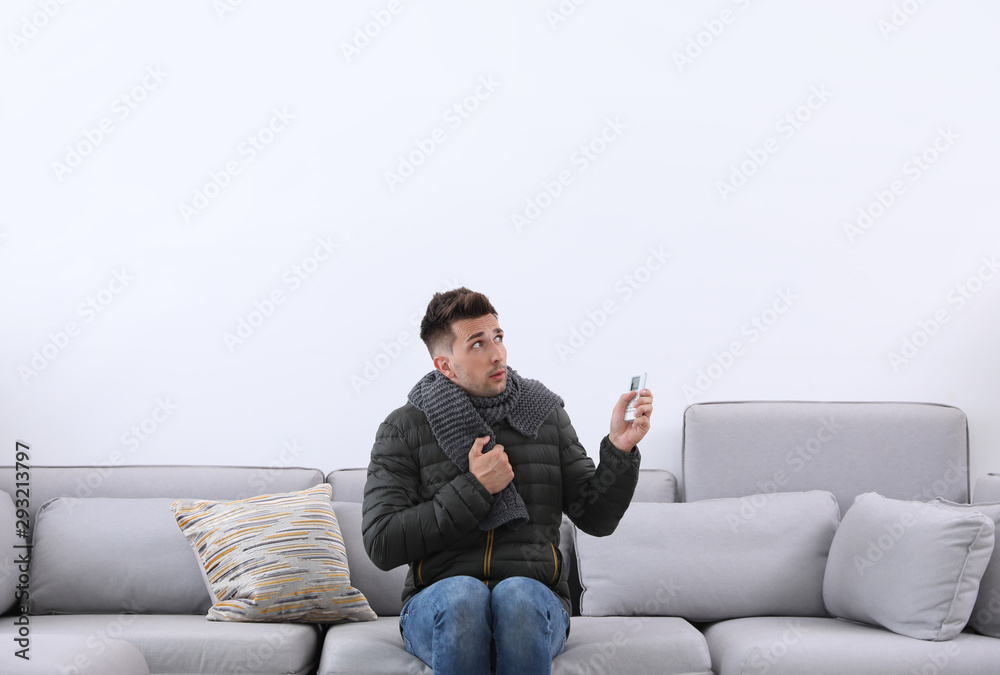 Young man with air conditioner remote control freezing on sofa at home