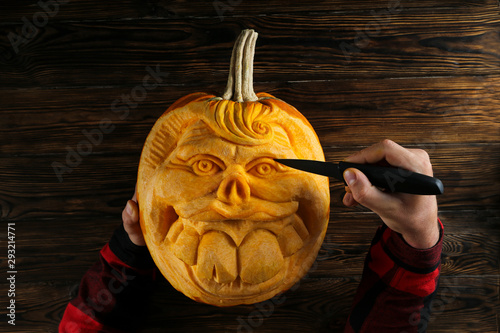 Top view shot of male hands carving creative human face shaped jack-o-lantern with big croocked teeth. Funny halloween pumpkin being carved by unrecognizable man. Close up, copy space, wood background photo