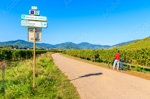 Sign with village names and directions and young woman biker among vineyards on road near Kientzheim village on Alsatian Wine Route, France