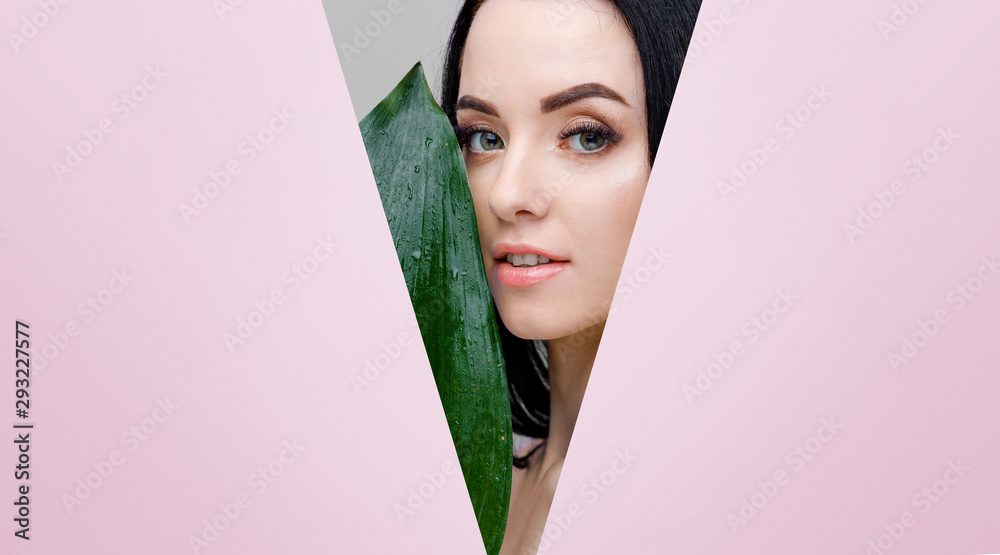 Portrait of young beautiful woman with healthy glow perfect smooth skin holds green tropical leaf. Model with natural nude make up look into the hole of pink paper. - obrazy, fototapety, plakaty 