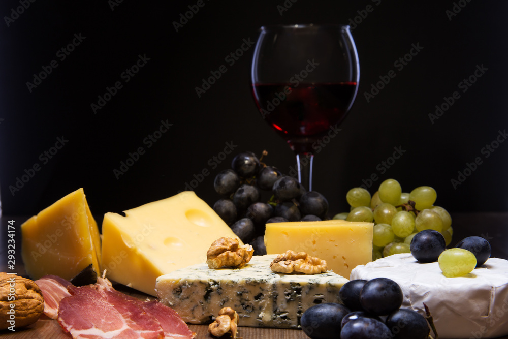 Assorted cheeses, nuts, grapes, fruits, smoked meat and a glass of wine on a serving table. Dark and Moody style. Assorted cheeses, nuts, grapes, fruits, smoked meat and a glass of wine on a serving table. Dark and Moody style.