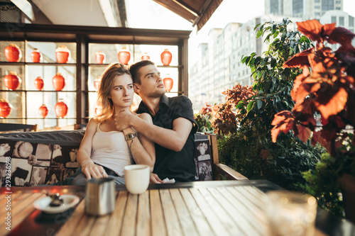 Portrait of young loving couple hugging on terrace in cafe with cup of coffee, looking away with serious face. Couple sitting on a date at a cozy cafe. Love story. © bodnarphoto