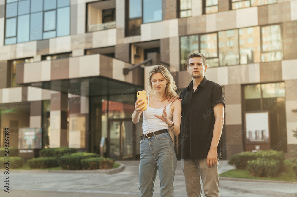 Street portrait of a fun couple in stylish casual clothes standing on the street and looking into the camera against the background of a modern apartment building.