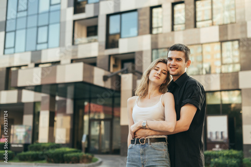 Positive Young family stands on the street against the backdrop of a modern apartment building,hugging and looking camera, wearing casual clothes.Street portrait of happy couple posing at camera
