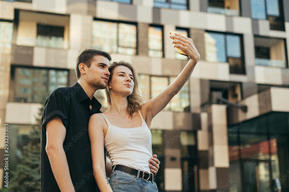 Beautiful young couple is standing outside on a summer day and taking selfie on a smartphone, looking at the camera and smiling, wearing stylish casual clothes.