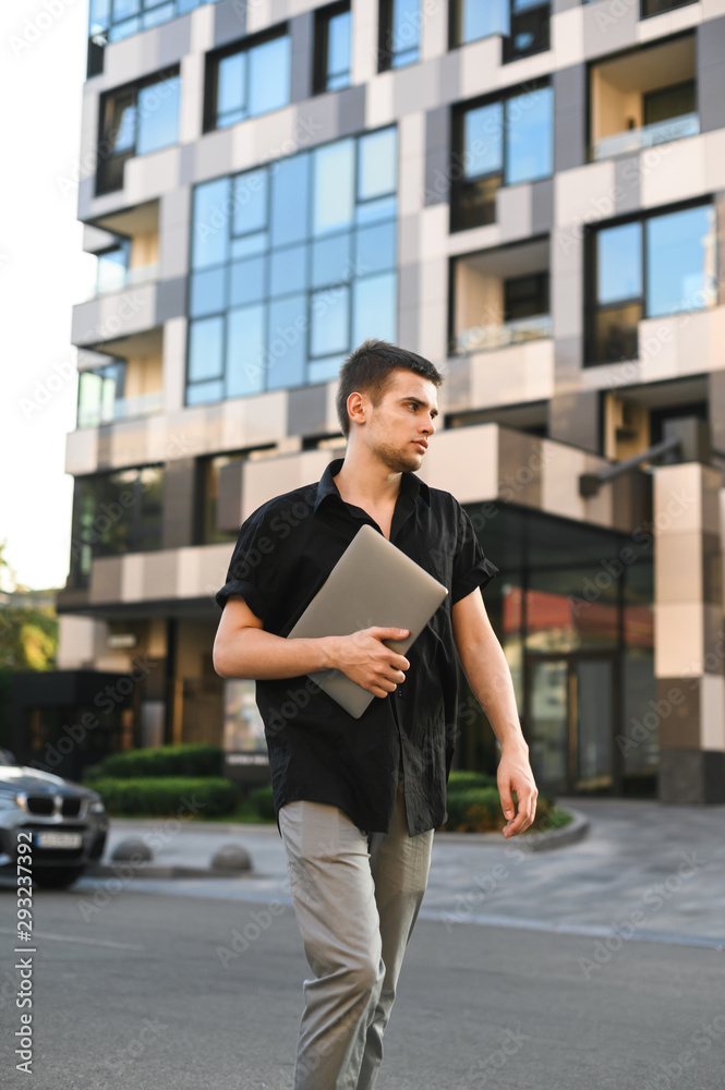 Fashionable student porter in casual clothes walking down the street with a laptop in his hand, wearing a black shirt and gray pants. Street style portrait of a stylish guy.