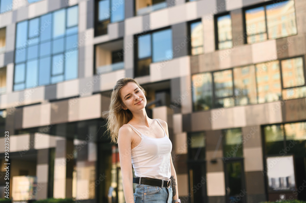 Smiling girl in a white T-shirt and jeans is standing outside, looking at the camera and smiling. Street portrait of stylish positive girl in casual clothes on a walk
