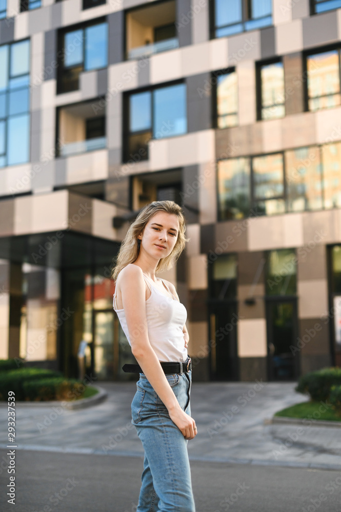 Portrait of attractive girl in casual clothes standing on the street against background of a modern building, looking into the camera, wearing jeans and a white T-shirt.Street vertical photo of girl