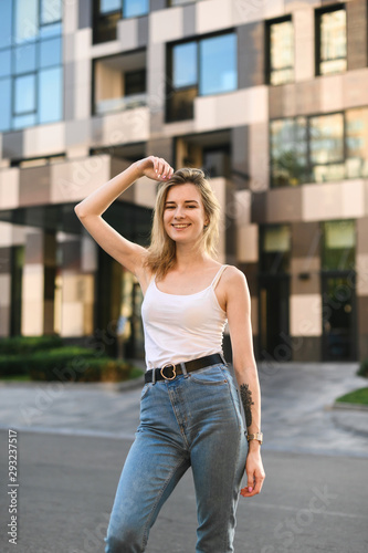 Street portrait of attractive girl in white tshirt and jeans standing on the street against the backdrop of modern architecture,posing at the camera and smiling,wearing stylish casual clothes.Vertical © bodnarphoto