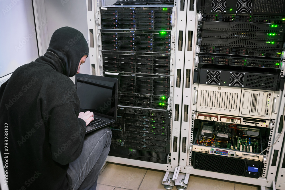 A hacker with laptop sits near the computer racks. View from the back. Criminal in black mask with datacents. Virus attack in server room. A hacker with laptop sits near the computer racks. View from the back. Criminal in black mask with datacents. Virus attack in server room.