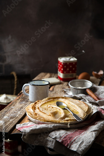 traditional russian or ukrainian stack of thin crepes with spoon of melted butter on top stands on rustic wooden table with vintage cup, strainer opposite concrete grey wall