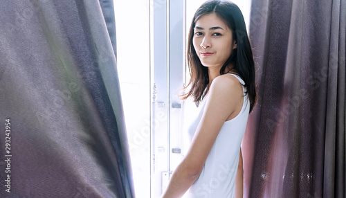 Enjoyment beautiful young woman slim fit body standing beside window in the morning while looking at camera hold curtain. Lifestyle relax concept.