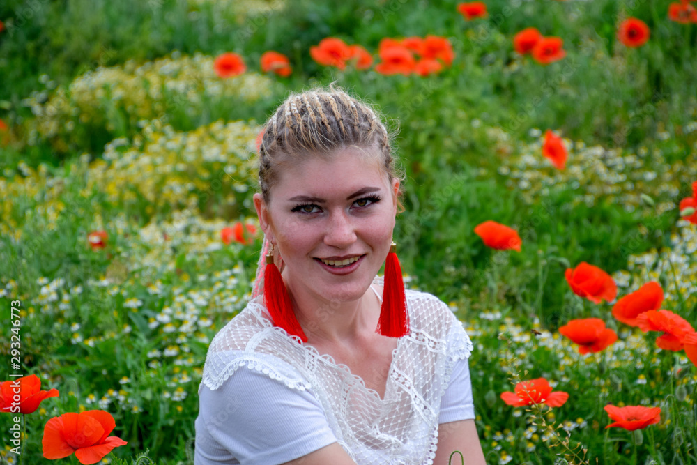 Blonde young woman in red skirt and white shirt, red earrings is in the middle of a poppy field. Blonde young woman in red skirt and white shirt, red earrings is in the middle of a poppy field.
