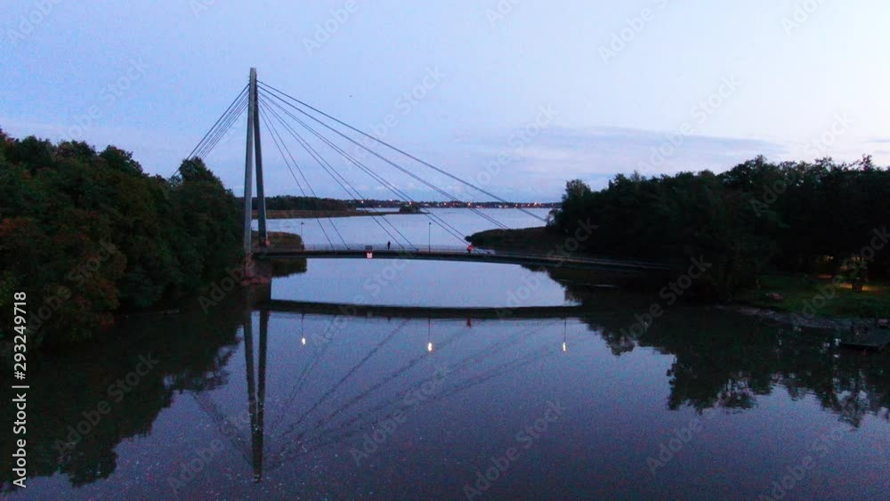 Crossing small bridge with metal wires. Calm river during evening with city lights in background. Crossing small bridge with metal wires. Calm river during evening with city lights in background.