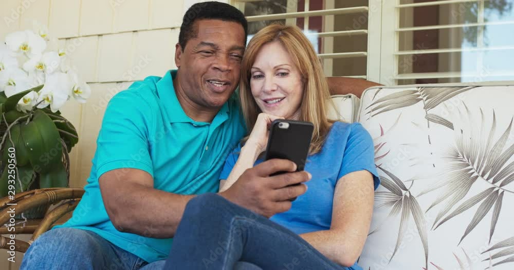 Happy older couple sitting on porch and using cell phone together. African American and Caucasian husband and wife looking at smartphone on sunny day outside home. Slow motion 4k handheld Happy older couple sitting on porch and using cell phone together. African American and Caucasian husband and wife looking at smartphone on sunny day outside home. Slow motion 4k handheld