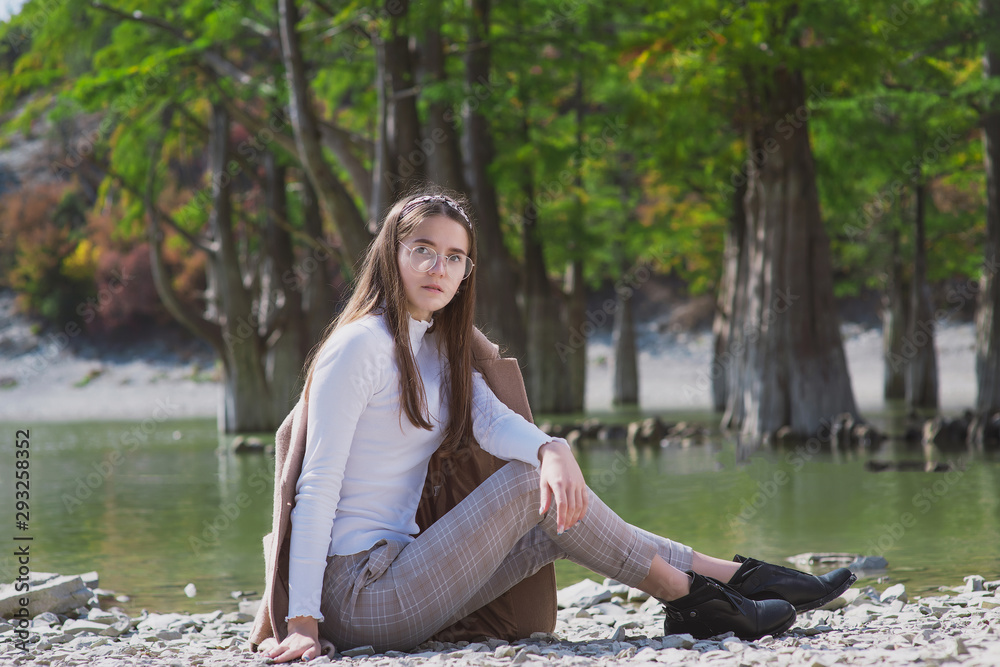 a beautiful, young brown-haired woman in a beige coat and a white turtleneck poses against a lake with cypresses in dark ankle boots