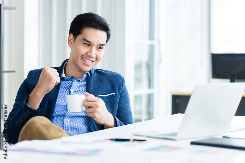 Happy of asian young businessman see a successful business plan on the laptop computer and holding a coffee cup, pen on wooden table background in office,business expressed confidence embolden Happy of asian young businessman see a successful business plan on the laptop computer and holding a coffee cup, pen on wooden table background in office,business expressed confidence embolden