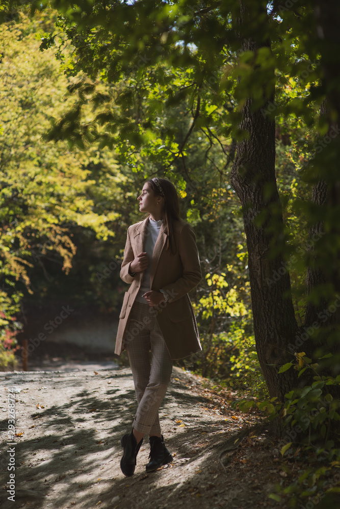 a beautiful, young brown-haired woman in a beige coat and a white turtleneck poses against the background of an autumn alley with yellow and red foliage a beautiful, young brown-haired woman in a beige coat and a white turtleneck poses against the background of an autumn alley with yellow and red foliage