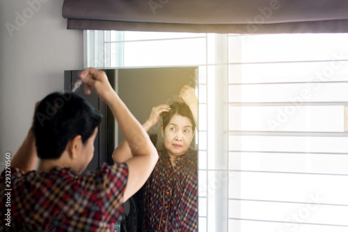 Elderly Asian woman plucking gray hair with tweezers on mirror