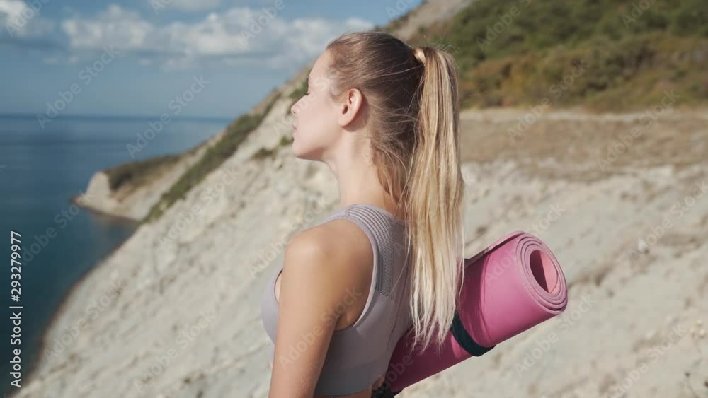 Woman holds yoga mat, looks at sea from cliff, breathes deeply, slow motion Woman holds yoga mat, looks at sea from cliff, breathes deeply, slow motion
