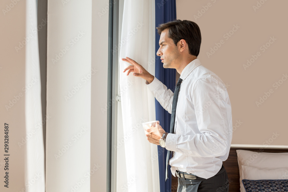 Handsome young businessman drinks coffee in front of the hotel window