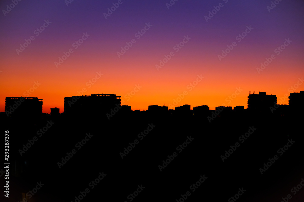Bright orange sunset over the city in the evening. Bright lights of high-rise buildings of urban buildings. Silhouette of construction city crane of building under construction