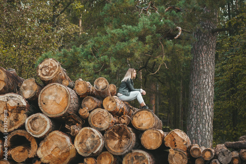 Beautiful woman in black leather jacket sitting on stack of large logs in autumn forest