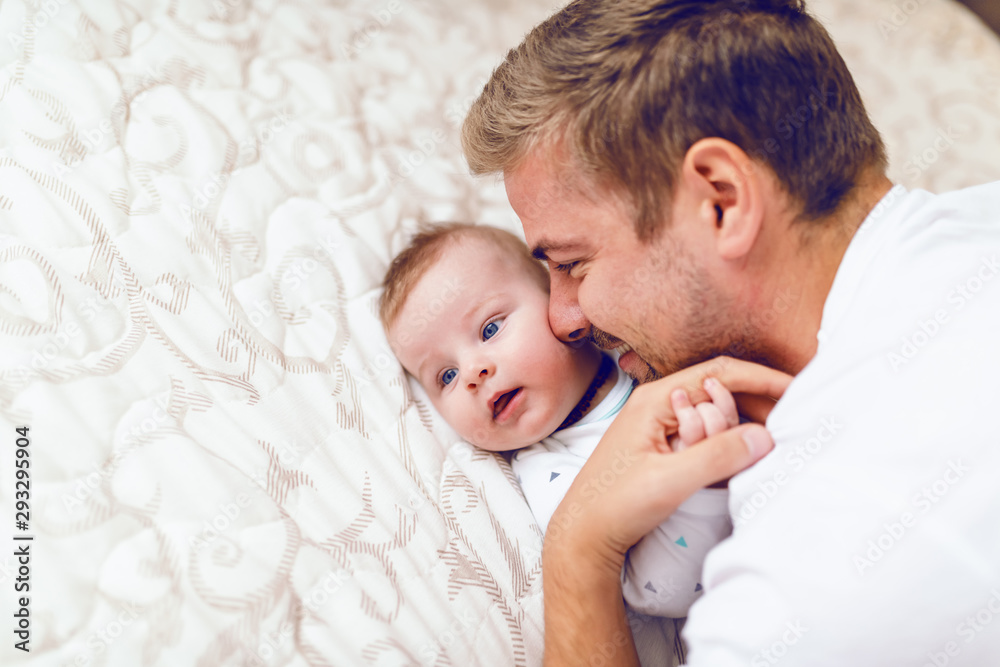 Close up of beautiful young caucasian dad cuddling with his six months old son. Baby lying in bed and hugging father. Close up of beautiful young caucasian dad cuddling with his six months old son. Baby lying in bed and hugging father.