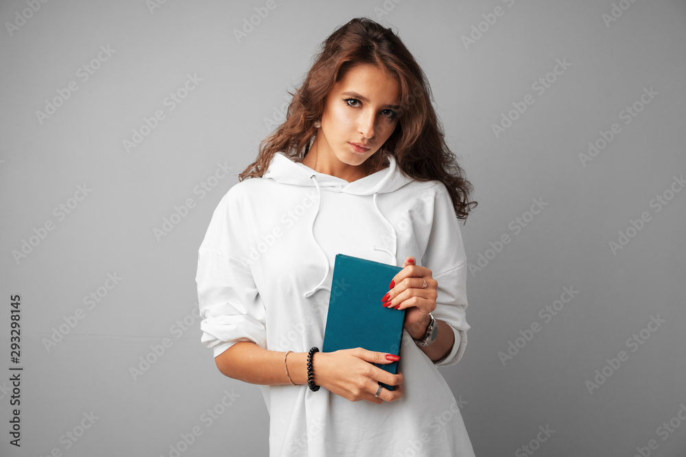 Fototapeta premium Student girl teenager in white hoodie holding a book in her hands over a grey background