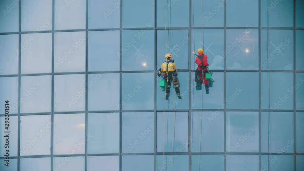 Two men workers in red and yellow work clothes cleaning the exterior blue windows of a skyscraper - industrial alpinism