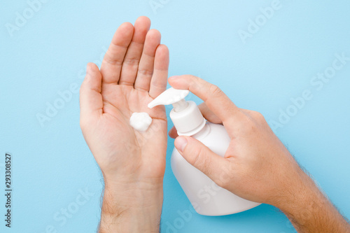 Perfect groomed man hands holding white bottle and using moisturizing cream on pastel blue background. Care about clean, soft and smooth body skin. Closeup.